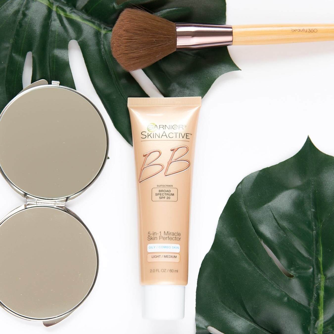 Garnier SkinActive BB Broad Spectrum SPF 20 5-in-1 Miracle Skin Perfector for Light/Medium on a white background next to two palm fronds, thick makeup brush, and a folding mirror.
